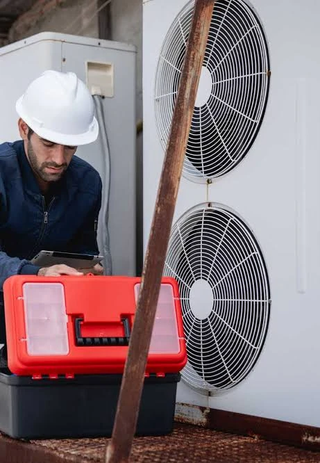 Technician wearing a white hard hat beside an outdoor HVAC unit and a red toolbox